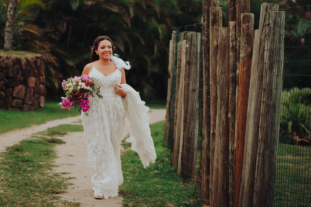 mulher com vestido de noiva com detalhe no ombro e segurando buquê com flores na cor branca e fúcsia