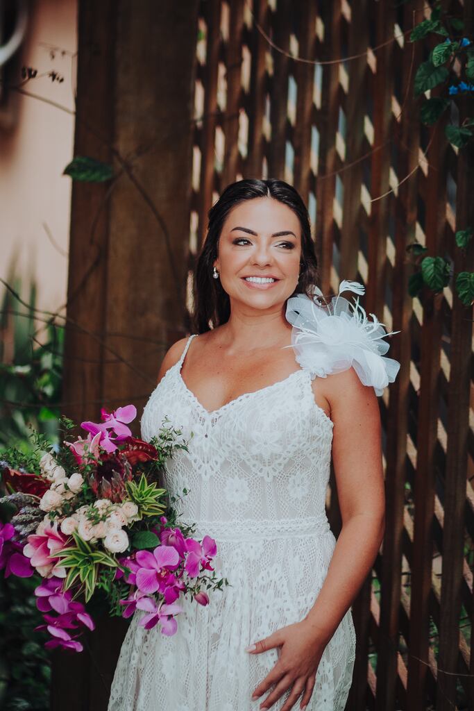 mulher com vestido de noiva com detalhe no ombr e segurando buquê com flores na cor branca e fúcsia