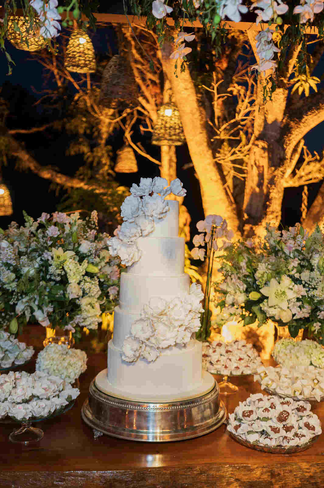mesa com bolo de casamento branco com flores com 5 andares ao lado de bandejas com doces
