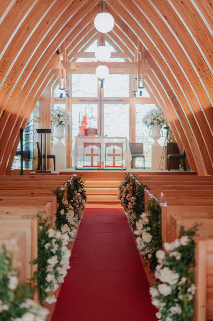caminho até o altar na igreja com tapete vermelho e bancos decorados com arranjos verdes e brancos