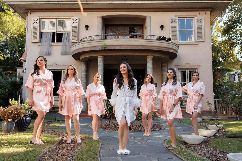 noivaa com robe branco ao lado de seis madrinhas com robes na cor rosa clara na frente de casa antiga
