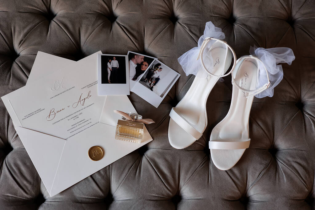 sandália branca com laço preso na lateral sobre o sofá cinza ao lado de convite branco e fotos polaroides