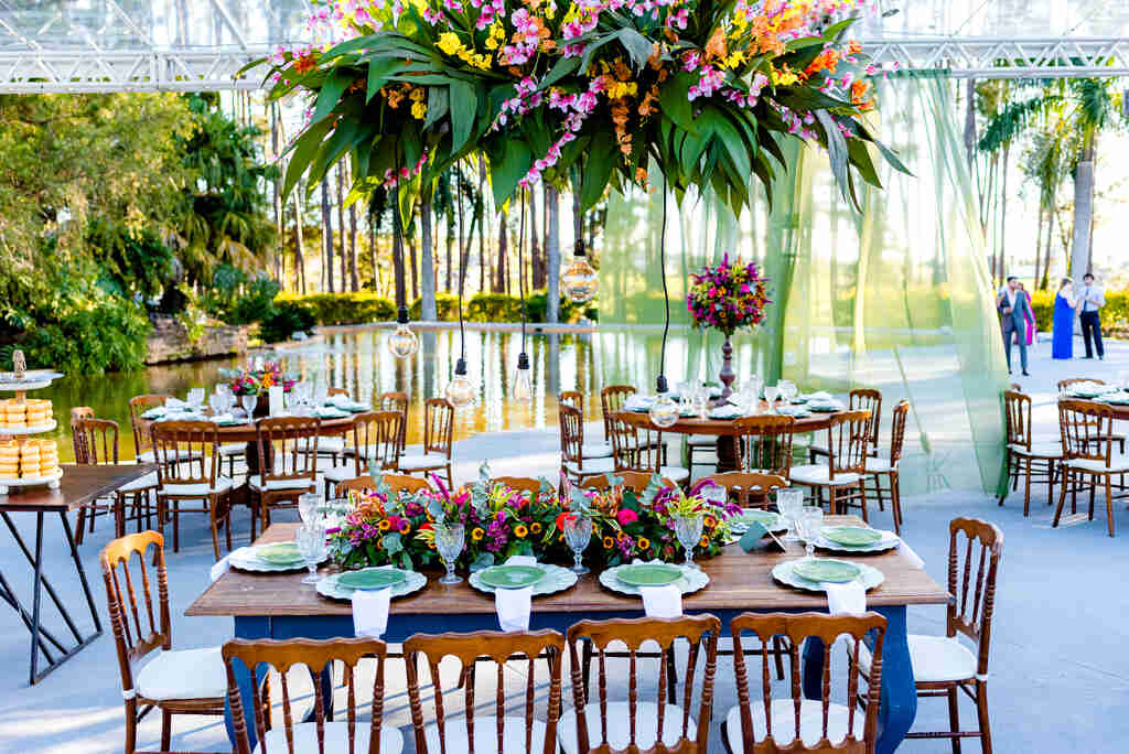 mesa retangular de madeira com mesa posta com vasos com flores rosas amarelas e roxas no centro arranjo pendente com flores coloridas acima da mesa