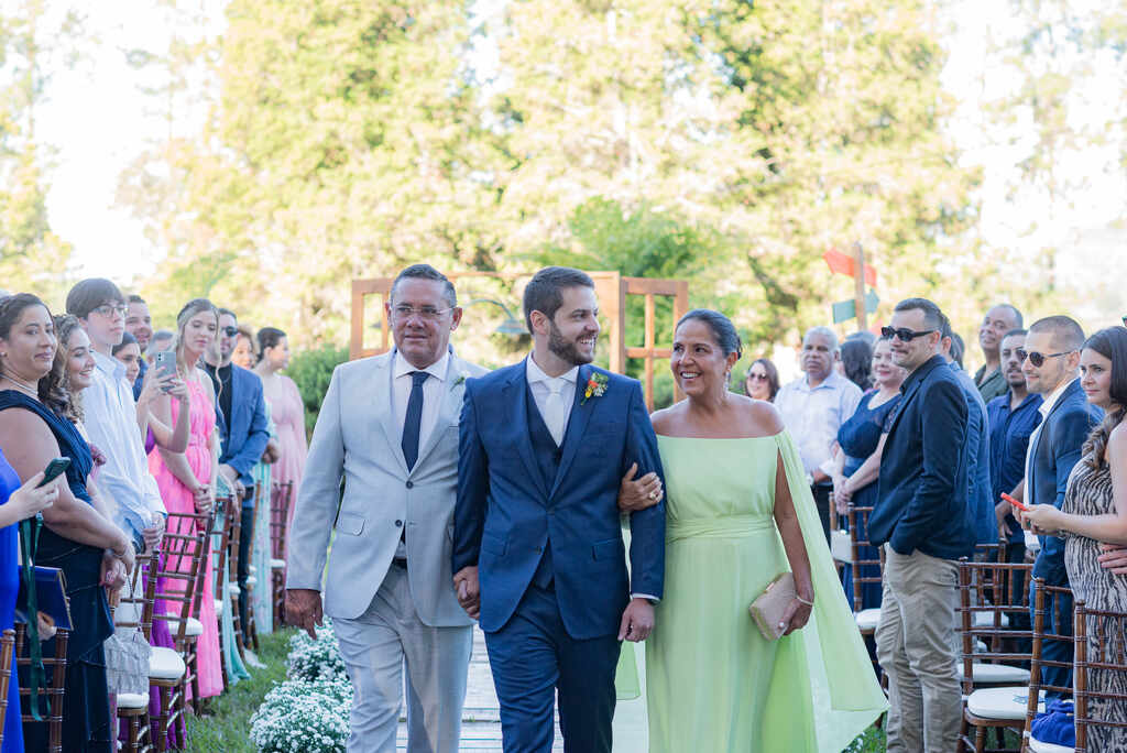 entrada do noivo com tenro azul ao lado do pai com terno cinza e mãe com vestido verde clarinho