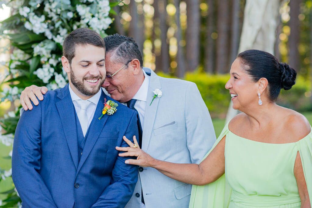 noivo com tenro azul sorrindo ao lado do pai com terno cinza e mãe com vestido verde clarinho
