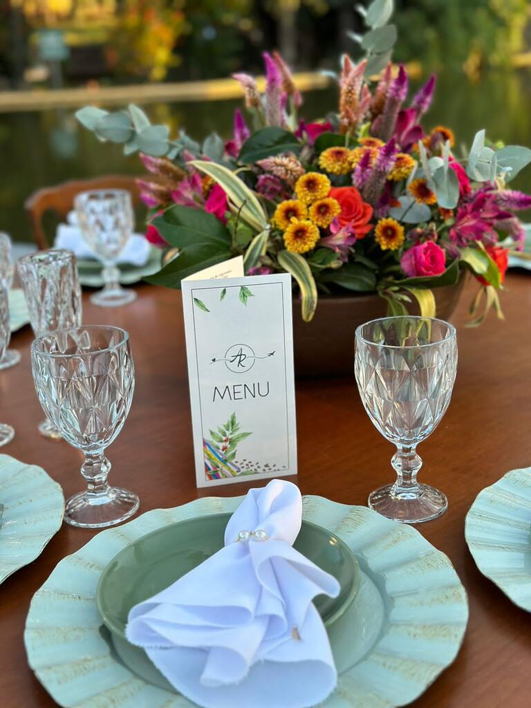 mesa posta com sousplat e prato verdes taças transparentes de vidro vasos com flores coloridas e menu com estampa de folhas