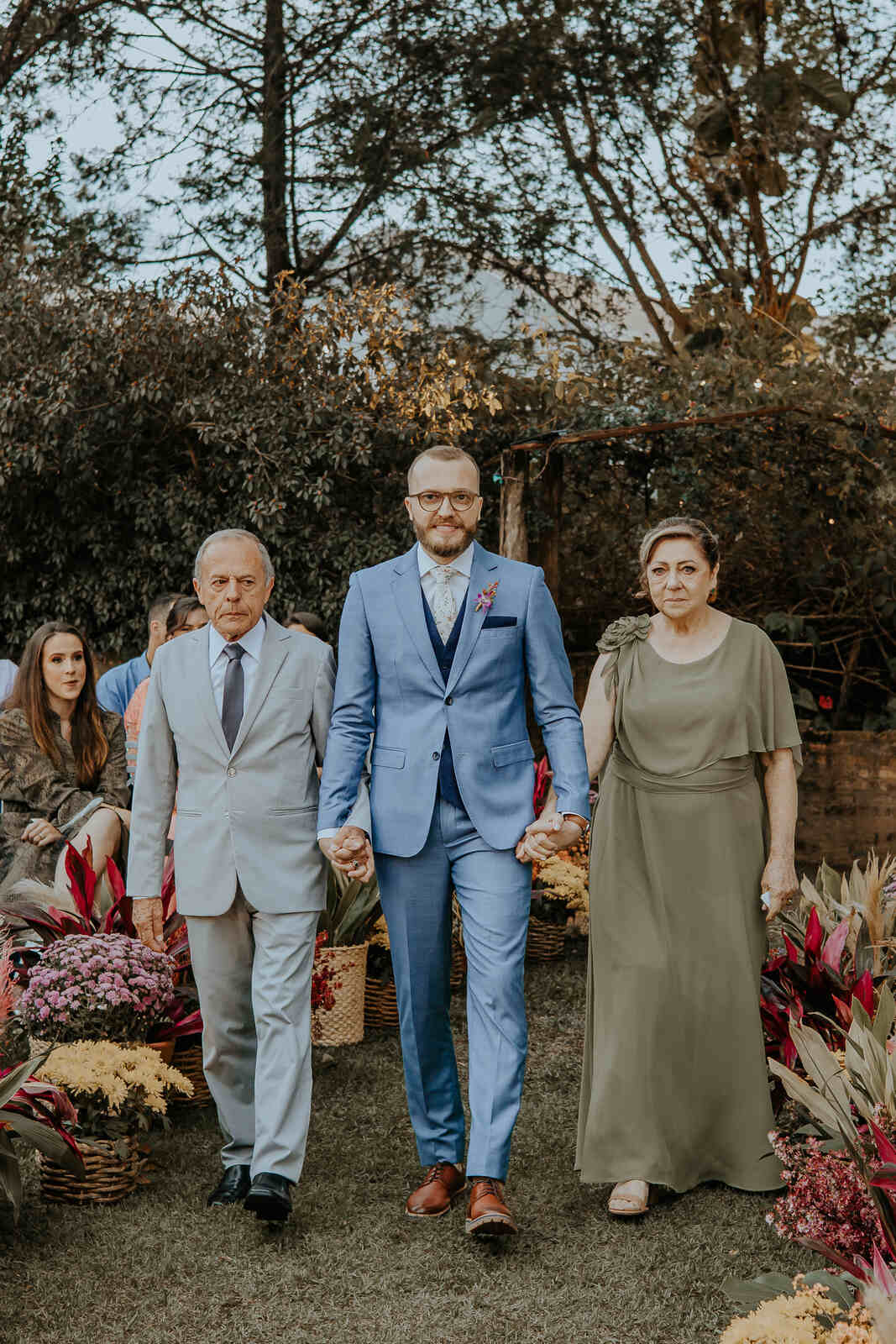 entrada do noivo com terno azul claro ao lado do pai com tenro cinza claro e mãe com vestido verde oliva