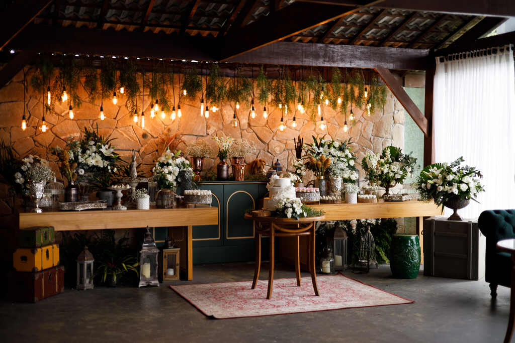 duas mesas de madeira com vasos de flores brancas ao lado de bandejas de doces e decorações e mesa central com mesa do bolo