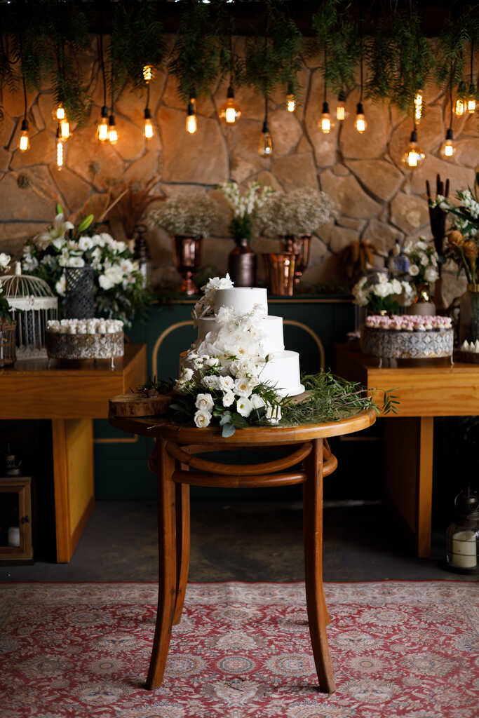 mesa redonda de madeira com bolo de casamento branco com três andares com flores brancas