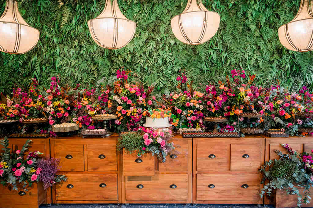 mesa de buffet madeira com bolo de casamento branco com flores no topo e ao lado bandejas com doces e arranjos com flores coloridas