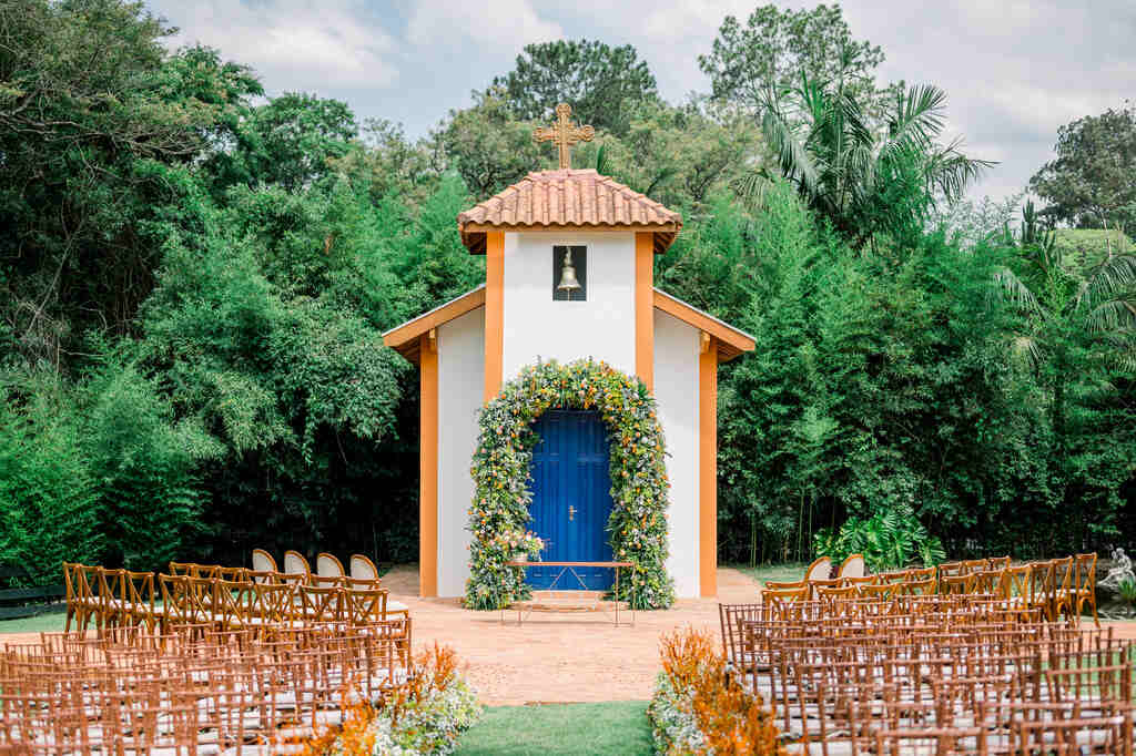 capela branca e laranja com porta azul e na frente altar com flores coloridas e cadeiras para assistir a cerimônia no campo