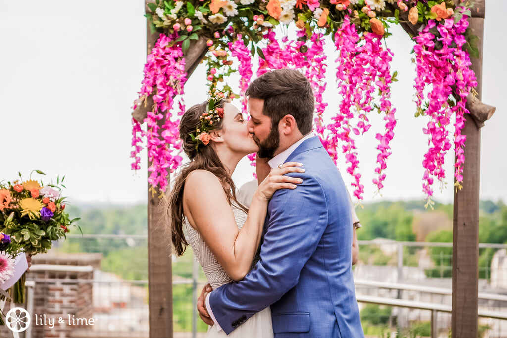 Noivo com terno azul beijando a noiva com coroa de flores sob altar rústico com decoração fucsia para casamento