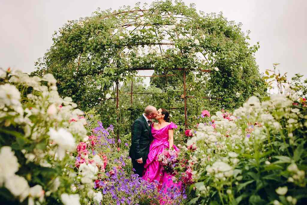 noiva com vestido na cor fucsia beijando o noivo com terno verde no jardim