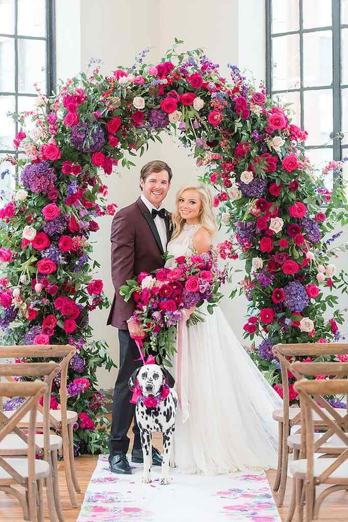 noivo com terno vinho ao lado de noiva com buquÊ com flores roxas e fucsia e cachorro dalmata ao lado dos noivos