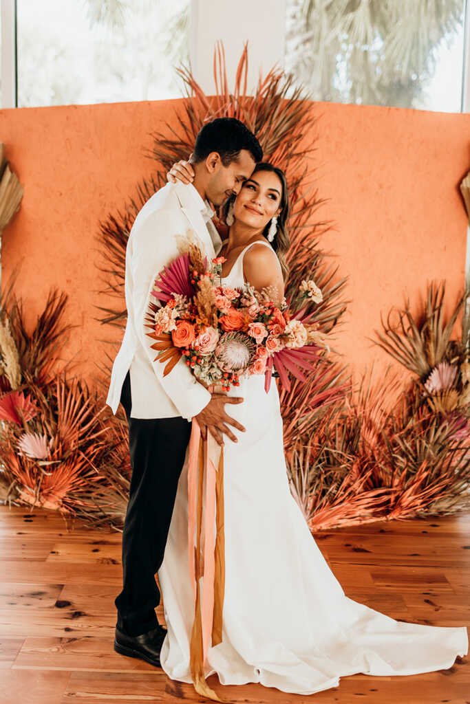 noivo com calça preta com terno branco abraçando noiva segurando buquê com flores laranja e rosê e ao fundo painel na cor coral
