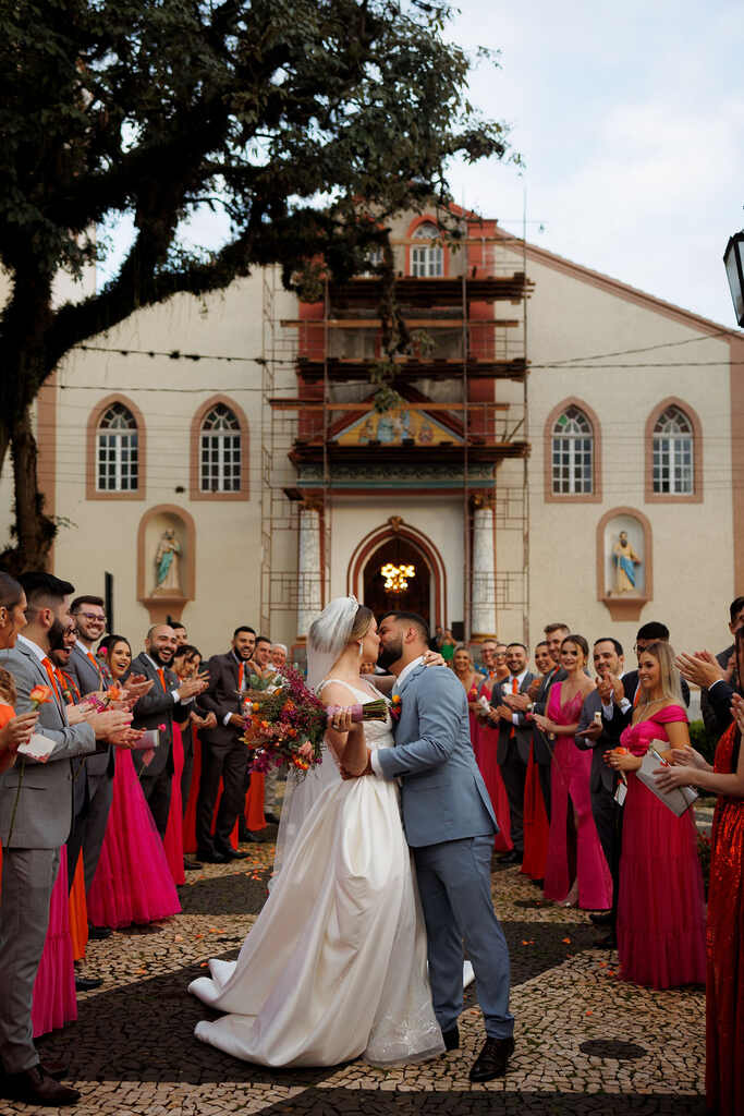 noivos se beijando na frente da igreja e ao lado madrinhas com vestidos rosa e laranja