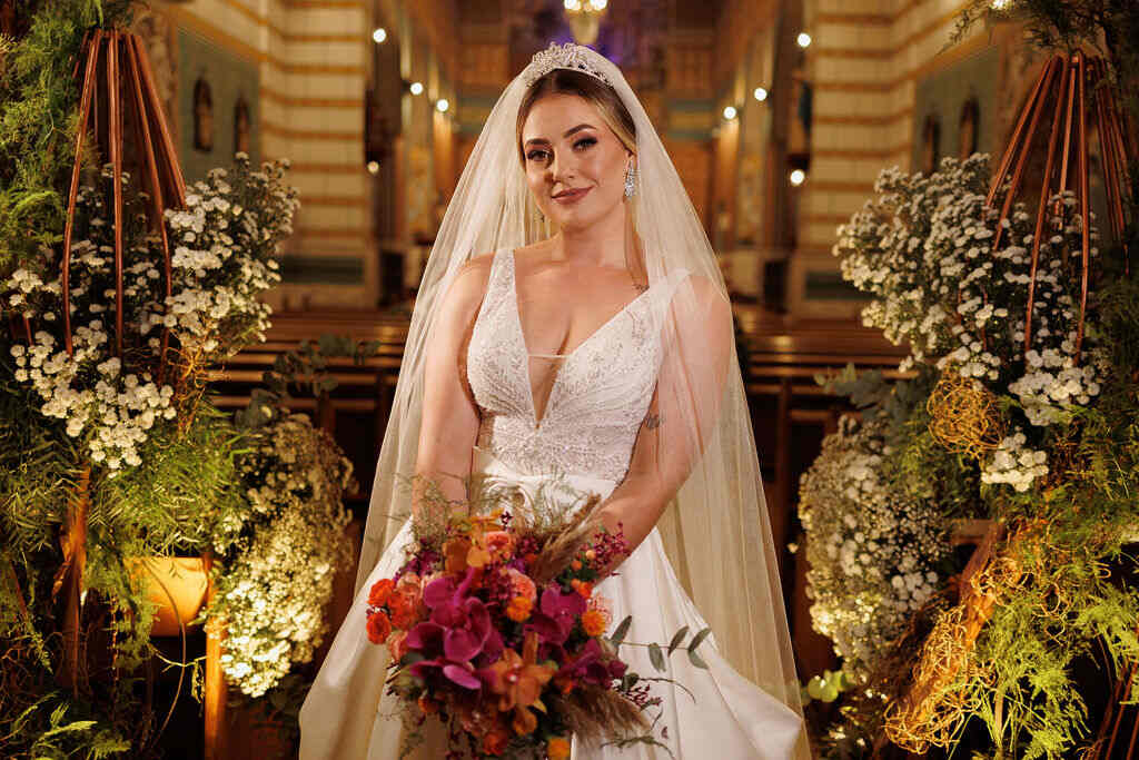 noiva com vestido com decote e véu longo segurando buquê com orquideas na cor fucsia e flores laranjas dentro da igreja decorada com plantas e flores brancas