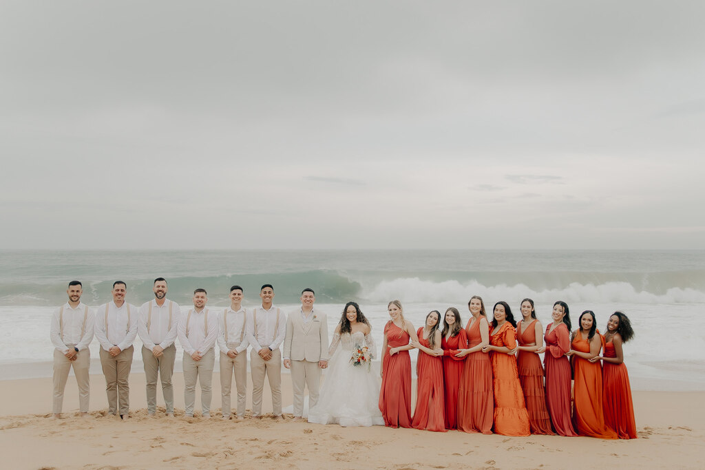 padrinhos com calça bege e supensório e camisa branca ao lado dos noivos e madrinhas com vestidos terracota na praia