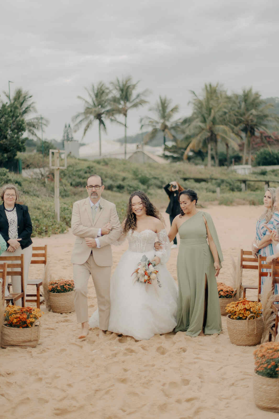 entrada da noiva ao lado do pai com terno bege e ao lado da mãe com vestido verde claro na praia
