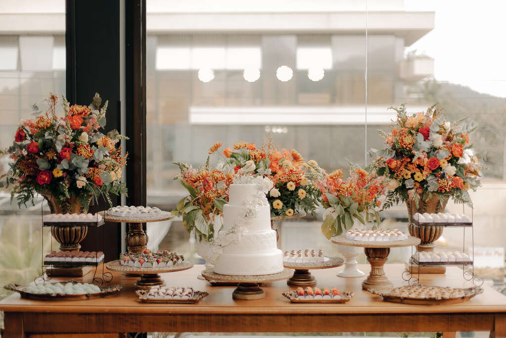 mesa de madeira com bolo de casmaento com quatro andares com flores brancas ao lado de bandejas com doces e vasos com flores vermelhas amarelas e laranjas ao fundo
