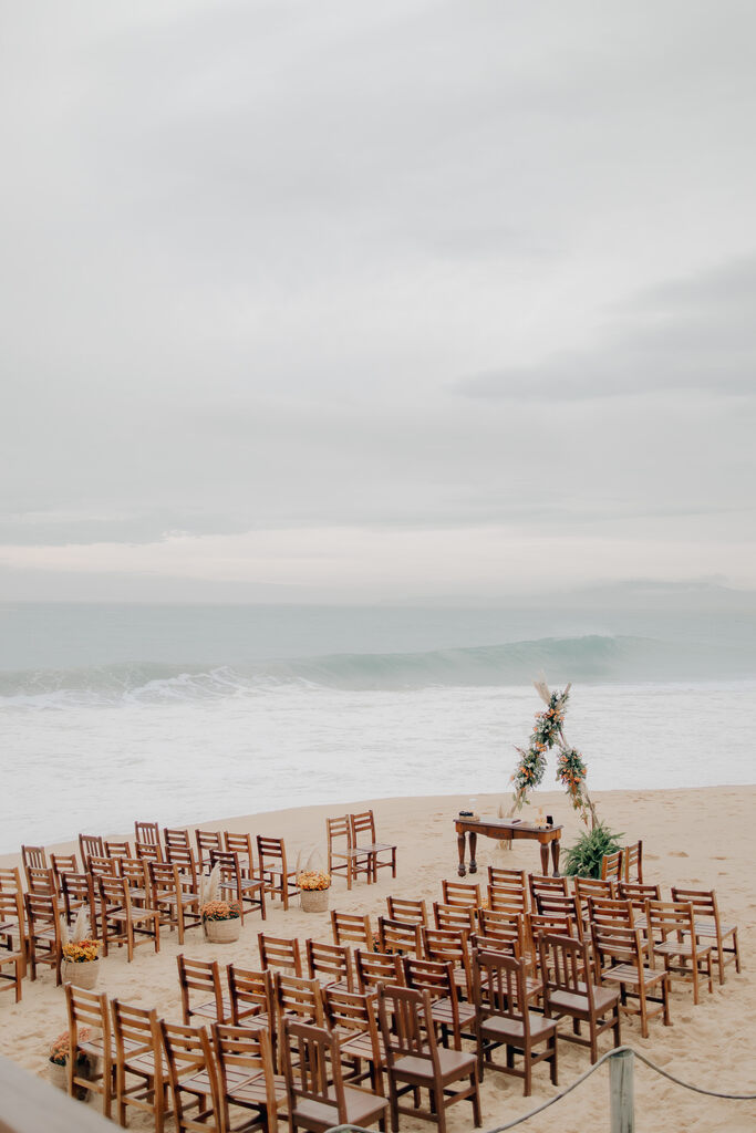 cadeiras de madeira escuras e altar triangular com capim dos pampas e flores brancas e laranjas na praia