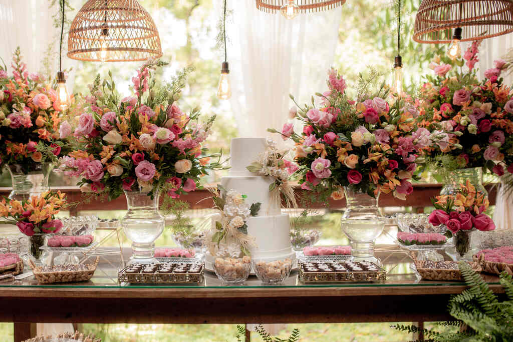 mesa de madeira com bandeja com doces e bolo de casamento branco com três andares ao lado de vasos de vidros com flores rosas amarelas e laranjas