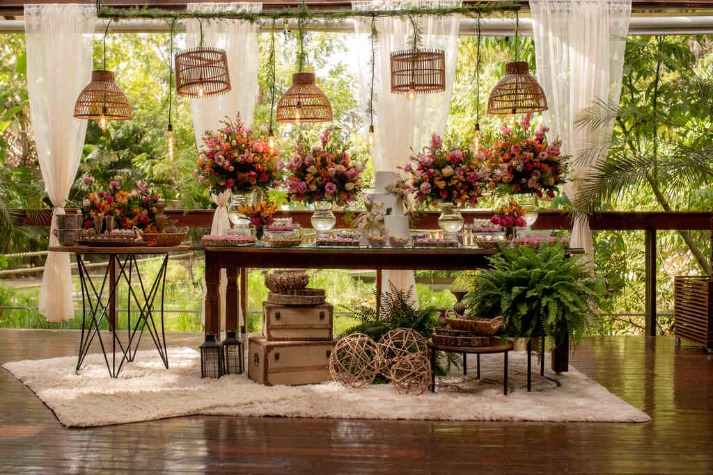 salão decorado com mesa de madeira com bolo de casamento ao lado de vasos de vidros com flores rosas amarelas e laranjas