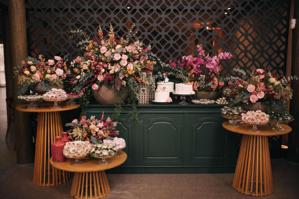mesa do bolo verde escura com trio de bolos de casamento e mesa decorada com bandejas com doces e arranjos com flores vermelhas rosas e na cor fucsia