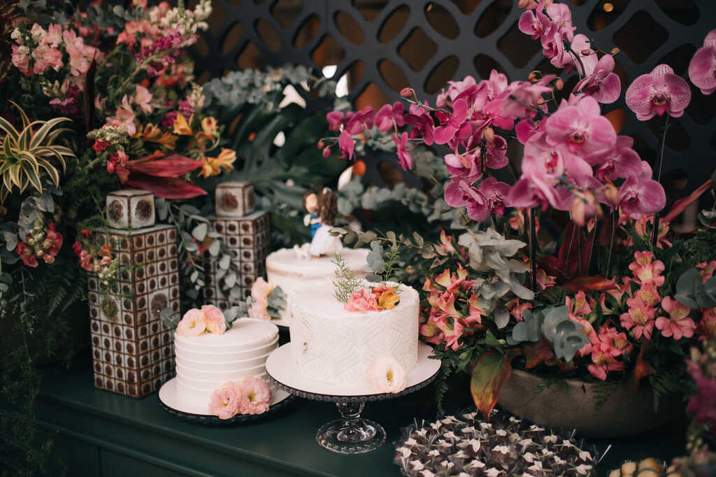 mesa do bolo verde escura com trio de bolos de casamento e mesa decorada com bandejas com doces e arranjos com flores vermelhas rosas e na cor fucsia