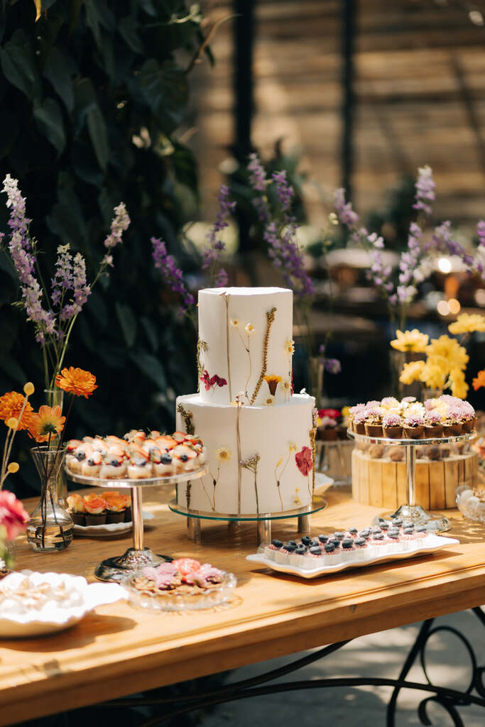 mesa de madeira com bolo de casamento com dois andares com flores prensadas e arranjos com flores lavandas cravos laranjas e flores amarelas