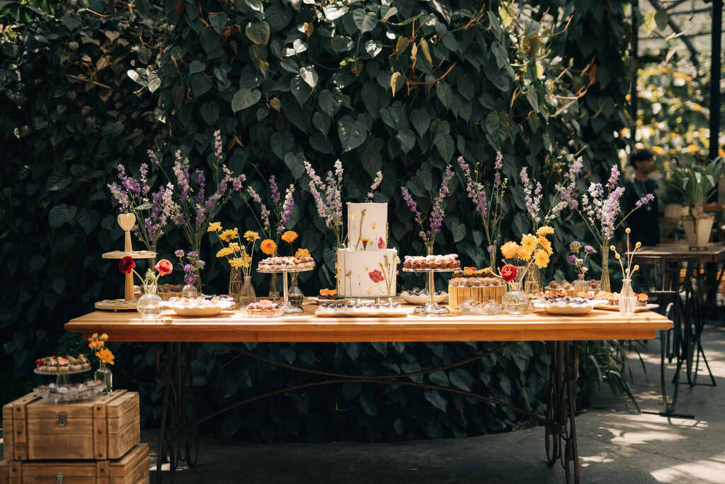 mesa de madeira com bolo de casamento com dois andares com flores prensadas e arranjos com flores lavandas cravos laranjas e flores amarelas