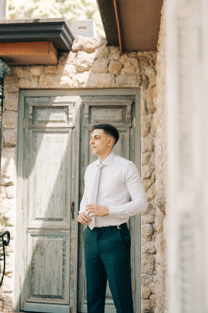 noivo com calça verde e camisa branca com gravata slim cinza clara na frente de porta antiga