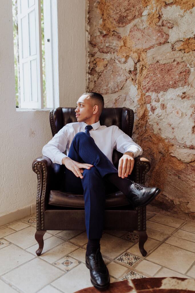 noivo com camisa branca e calça azul marinho sentado em poltrona marrom de couro em sala rústica