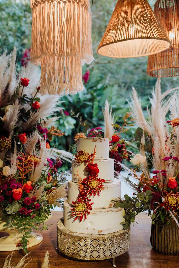mesa de madeira rústica ao ar livre com bolo de casamento com vasos com capim dos pampas e flores e luminarias no estilo boho
