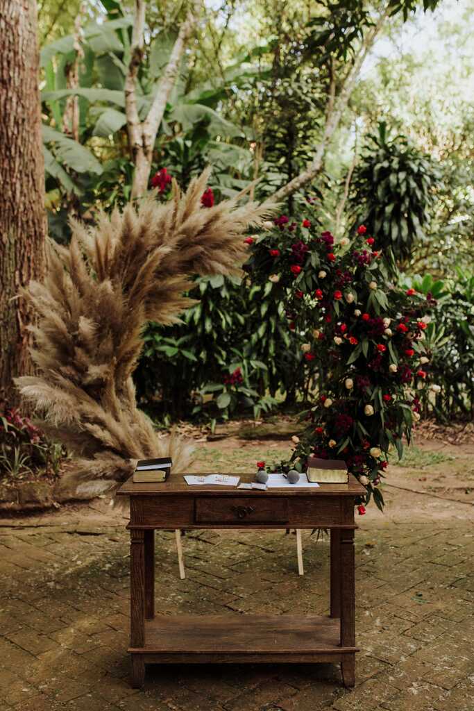 mesa de madeira rústica na frente de altar reondo com capim dos pampsa e metade com flores vermelhas ao ar livre