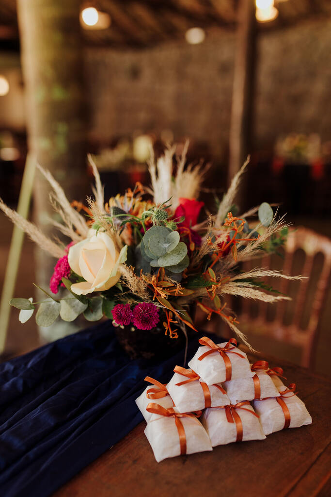 pilha de bem-casados com embalagens brancas com fita laranja ao lado de tecido azul escuro e vaso com flores brancas e rosas com capim dos pampas