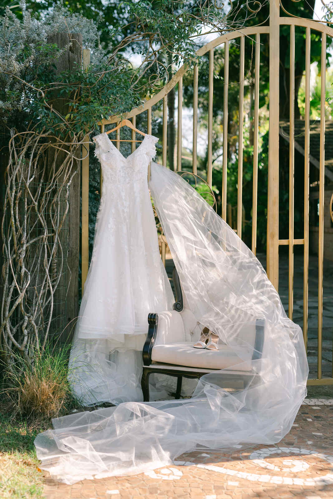 vetsido de noiva e véu longo pendurado no portão vintage ao lado de poltrona clássica com sandália em cima