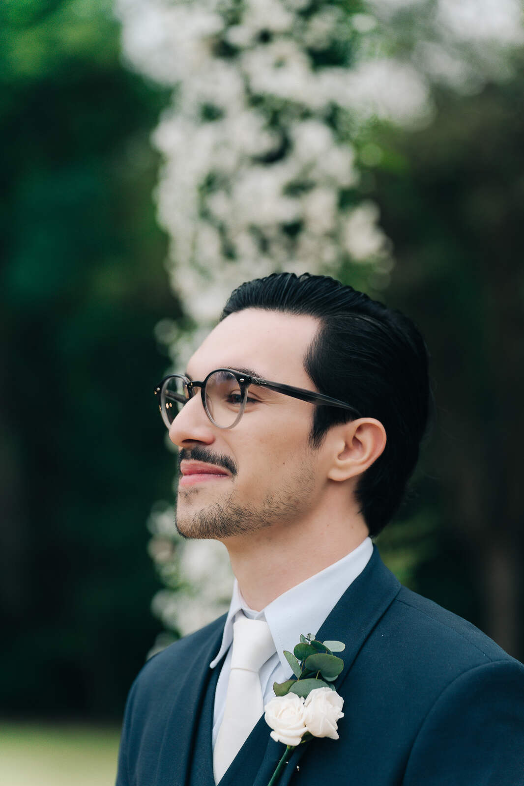 noivo com cabelo penteado para trás camisa e gravata branca e óculos e terno azul escuro aguardando a noiva no altar