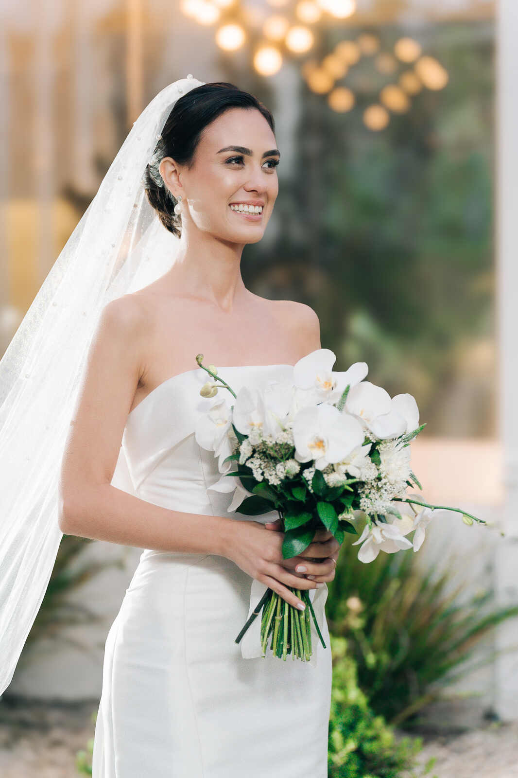 noiva com vetsido minimalista segurando buquê com flores brancas e orquideas e véu com aplicação de pérolas 
