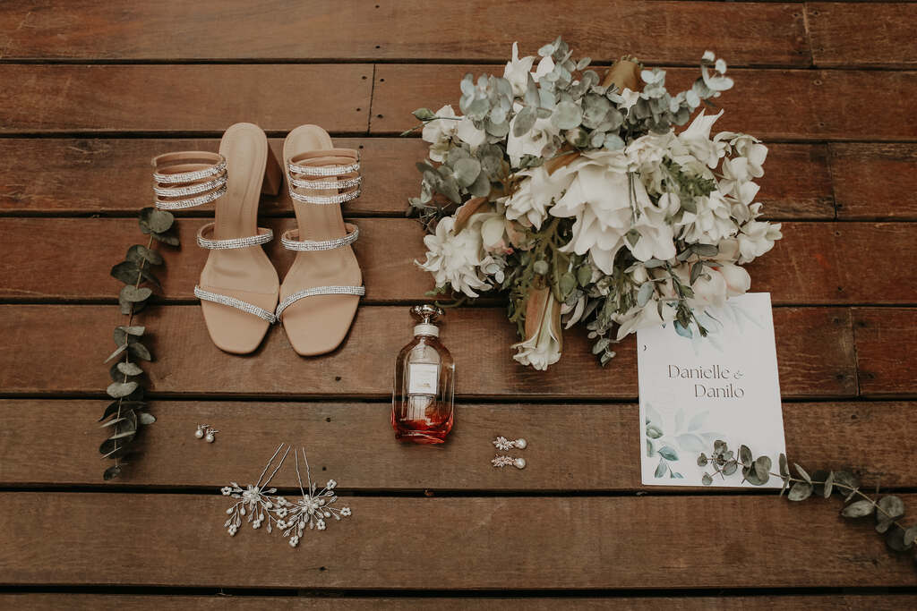 sandália com detalhes prateados ao lado do buquê com flores brancas vidro de perfume e convite de casamento branco com estampa com folhas