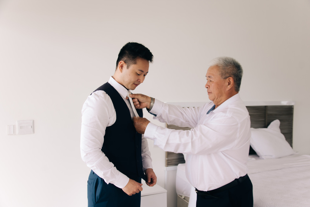 pai do noivo com camisa branca ajustando gravata do noivo dentro do quarto