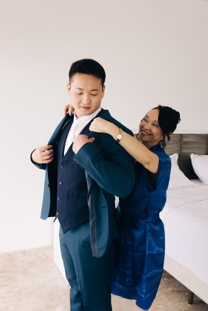 mãe do noivo com robe azul escuro ajustando o terno do noivo dentro do quarto