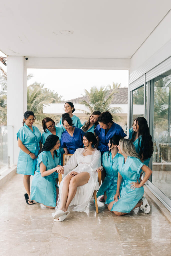 noiva com robe branco sentada na cadeira e madrinhas com robes na cor azul na varanda