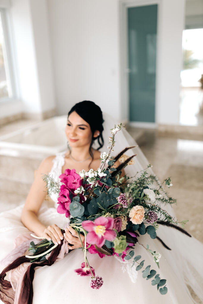 mulher com vetsido de noiva com saia bufante cabelo preso e véu longo segurando buquê desconstruido com flores rosas brancas e roxas sentada na cadeira