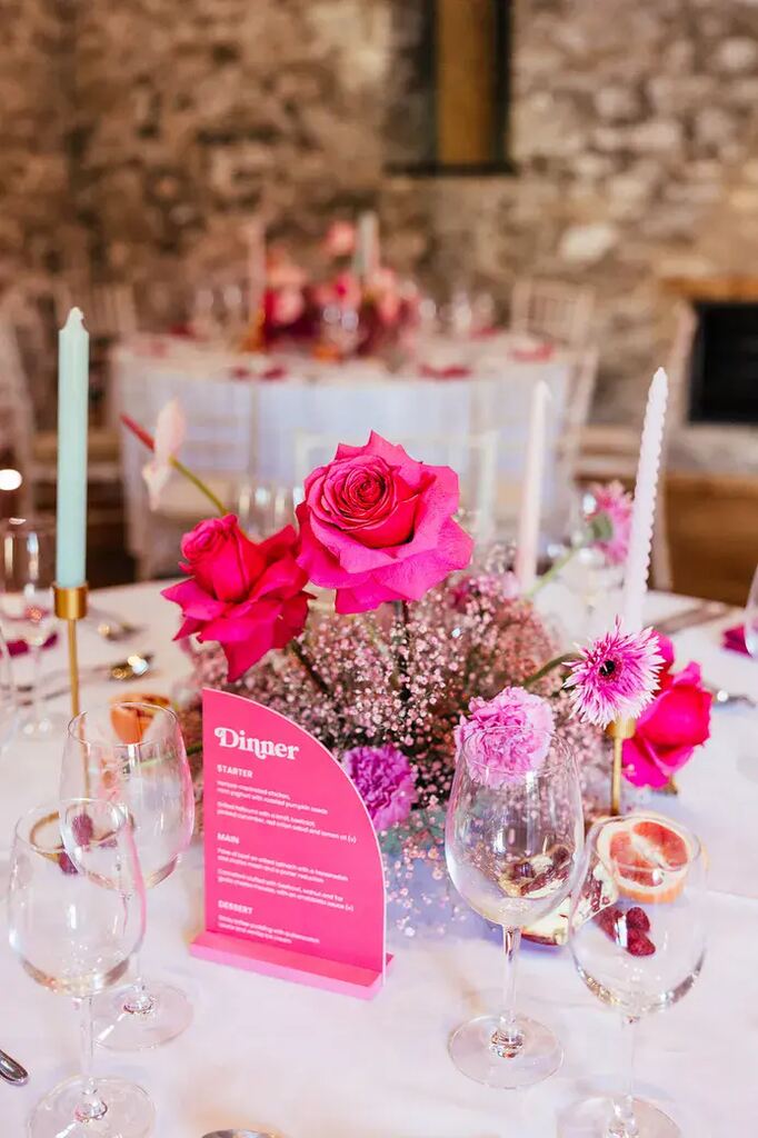 mesa posta com toalha branca flres no centro velas coloridas taças e marcador de mesa na cor pink