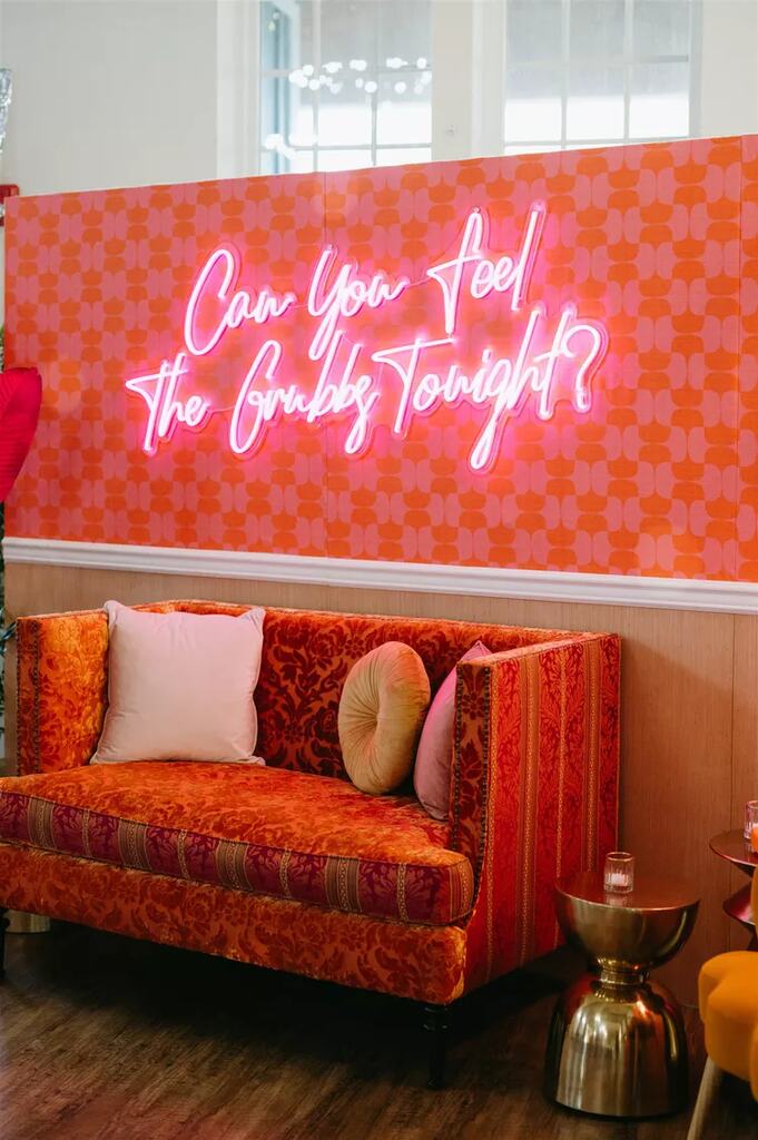 lounge com sofá com estampa étnica laranja e rosa e ao fundo papel de parede quadriculada e letreiro neon pink