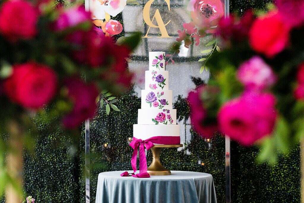 mesa com toalha azul com bolo de casamento branco com cinco andares com estampa de flores roxas