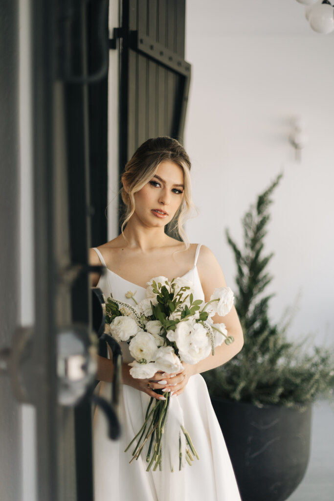 noiva com vestido minimalista de alças finas e cabelo preso com duas mechas soltas com batom nude segurando buquê com flores brancas
