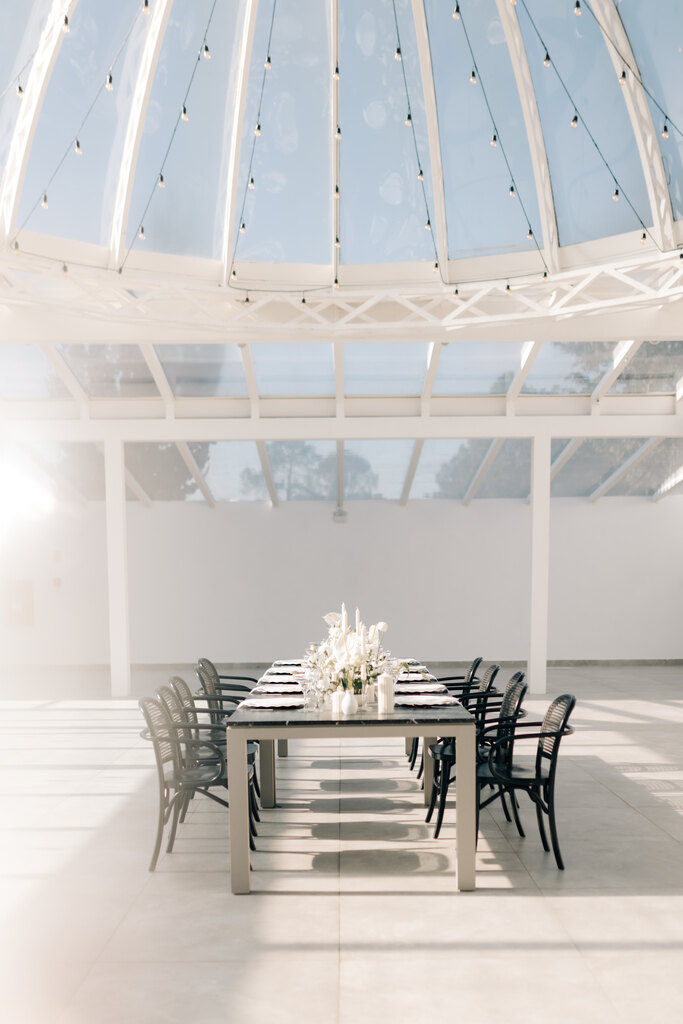 mesa comprida com cadeiras pretas e flores brancas no centro dentro de salão branco e minimalista com janelas de vidro