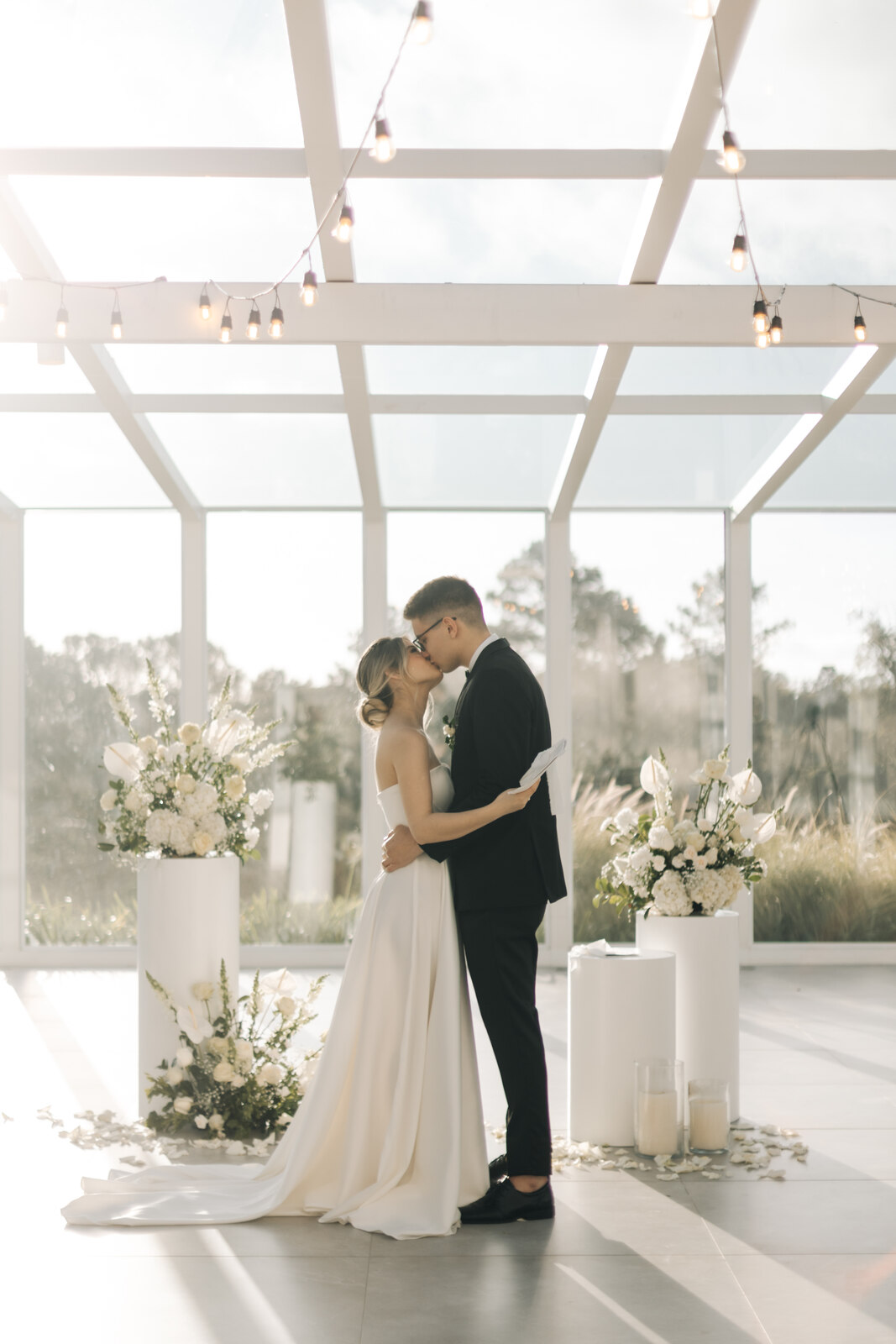 casal se beijando no altar dentro de salão branco minimalista com janelas e cobertura de vidro